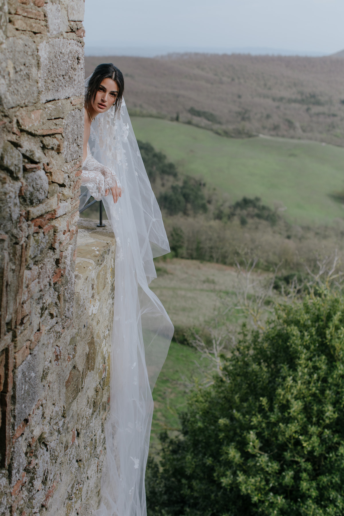 A bride in a white dress and veil leans out from a stone wall, overlooking a rural landscape with hills and greenery.