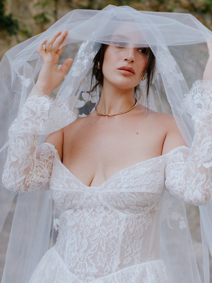 A bride in a white lace wedding dress and veil stands outdoors, holding the veil with both hands and looking at the camera.
