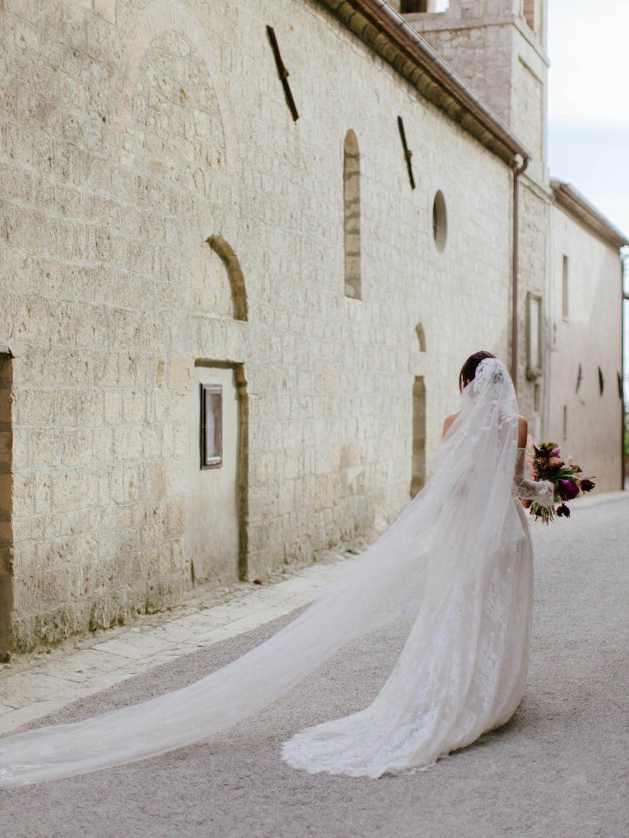 A bride in a long white dress and veil walks alone down a narrow stone street, holding a bouquet of flowers.