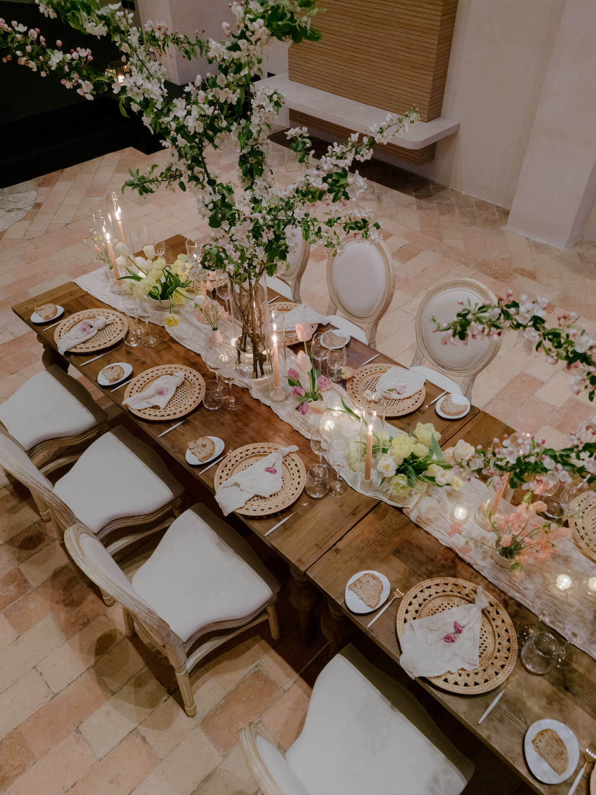 A long wooden table set for a formal meal with elegant plates, napkins, and floral centerpieces, viewed from above.