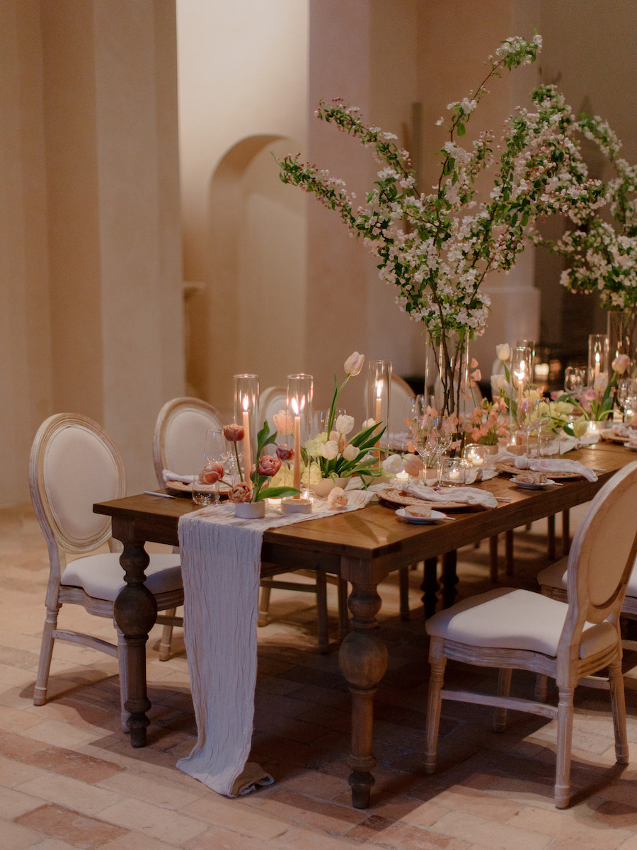 Elegant dining table set with candles, glassware, plates, and tall floral arrangements, surrounded by cushioned chairs in a softly lit room.
