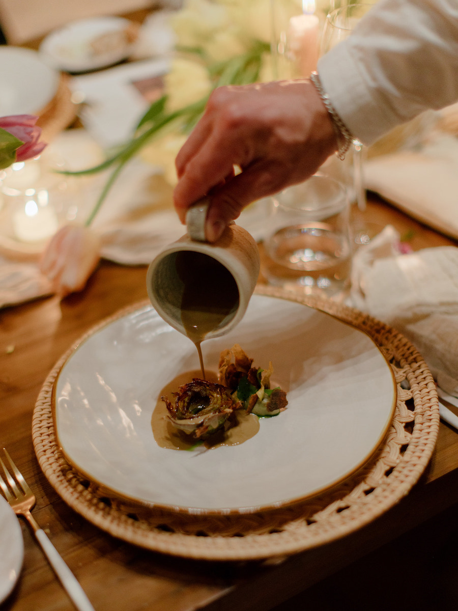 A hand pours sauce from a small pitcher onto a plated dish with a garnish, set on a woven placemat at a decorated dining table.