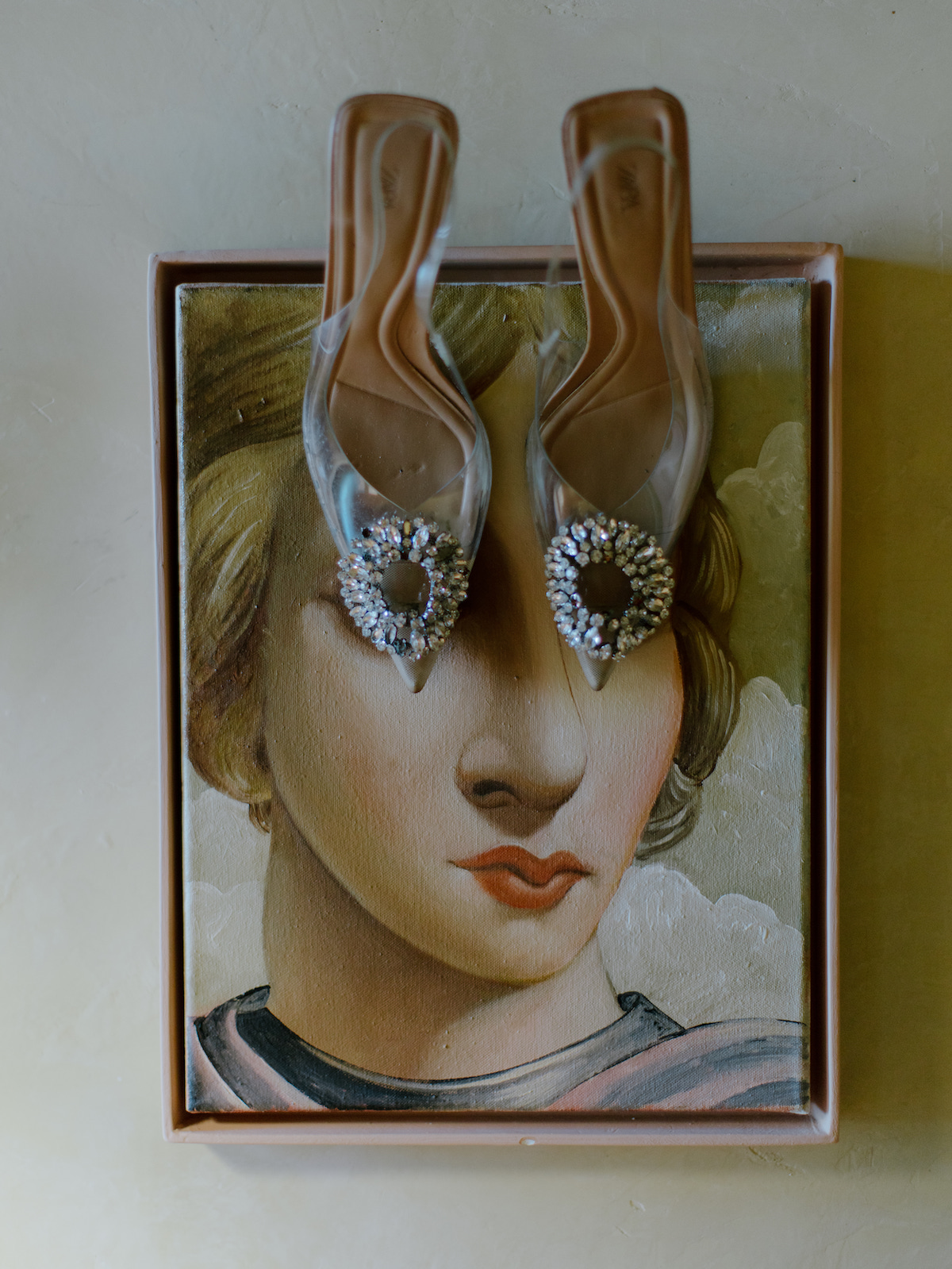 A painting of a woman’s face with two jeweled shoes placed on top, aligned to cover her eyes.