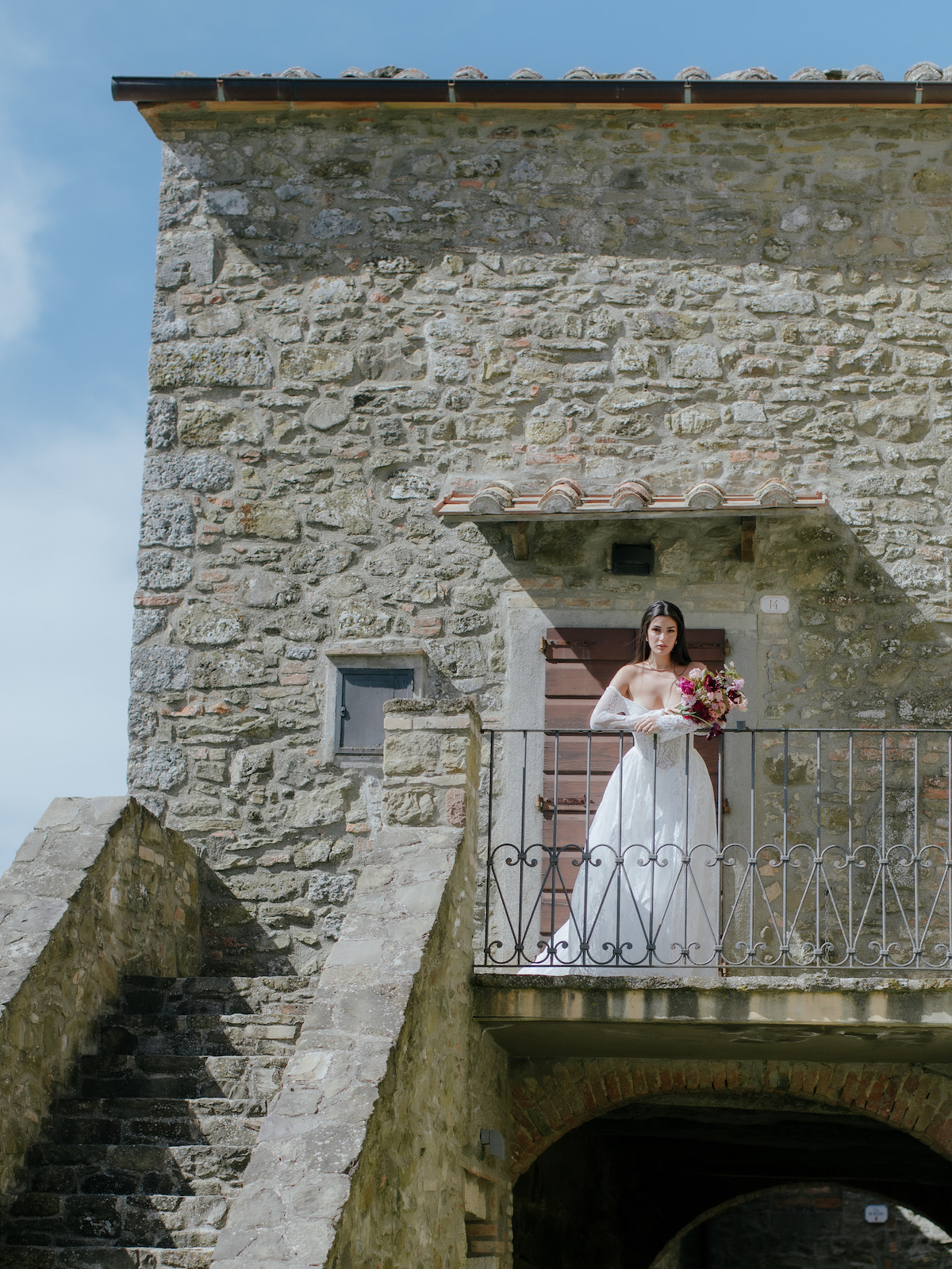 A bride in a white dress stands on a stone balcony holding a bouquet, with a stone building and staircase in the background.