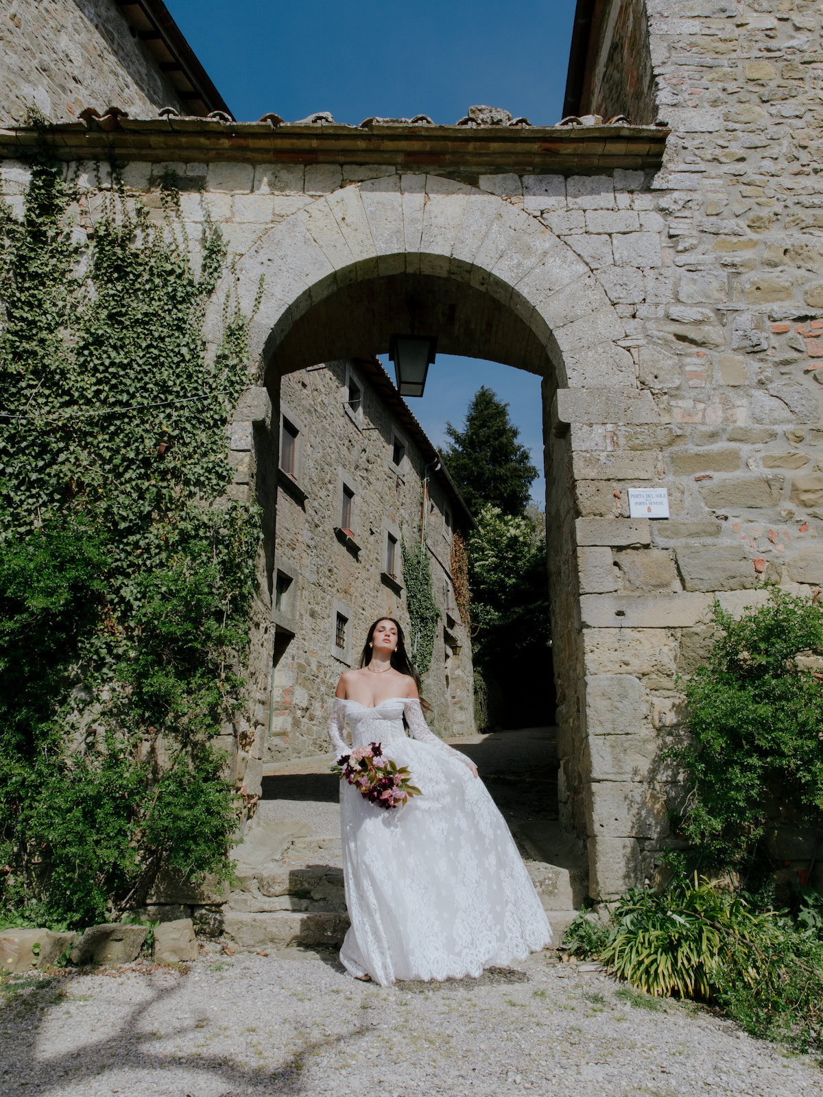 A bride in a white wedding dress stands holding a bouquet under a stone archway, surrounded by old stone buildings and greenery.
