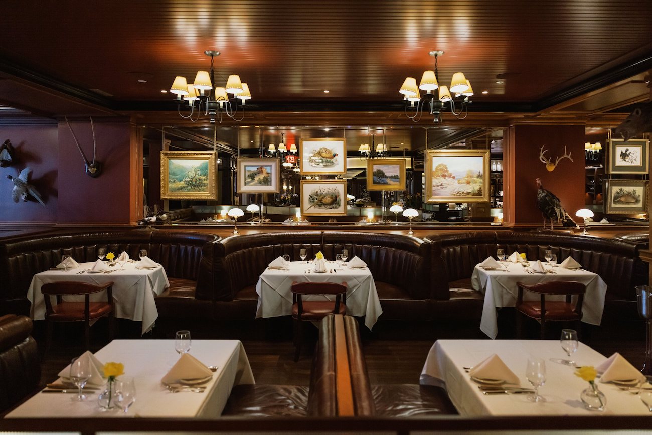 A dimly lit restaurant with dark leather booths, white tablecloths, framed art on the walls, and chandeliers hanging from a wooden ceiling.