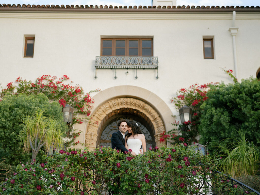 A couple dressed formally stands close together on a balcony in front of a large arched doorway, surrounded by flowering plants and greenery.