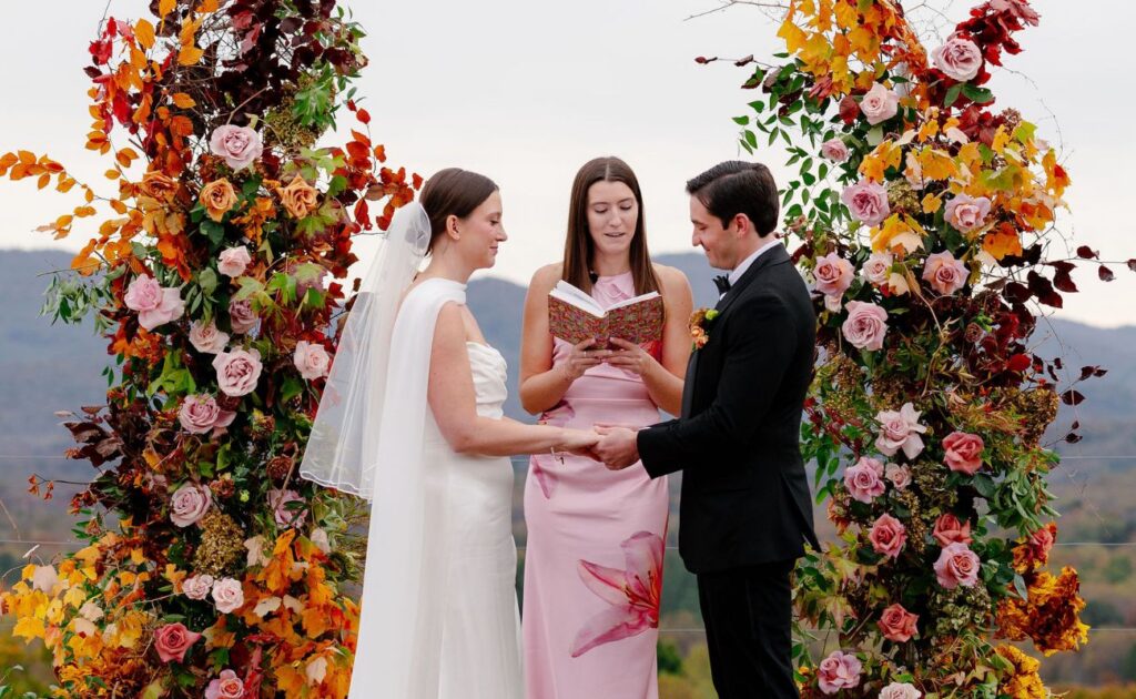 A bride and groom hold hands during an outdoor wedding ceremony, standing under a floral arch with an officiant reading from a book.