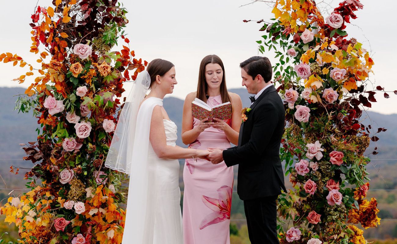 A bride and groom hold hands during an outdoor wedding ceremony, standing under a floral arch with an officiant reading from a book.