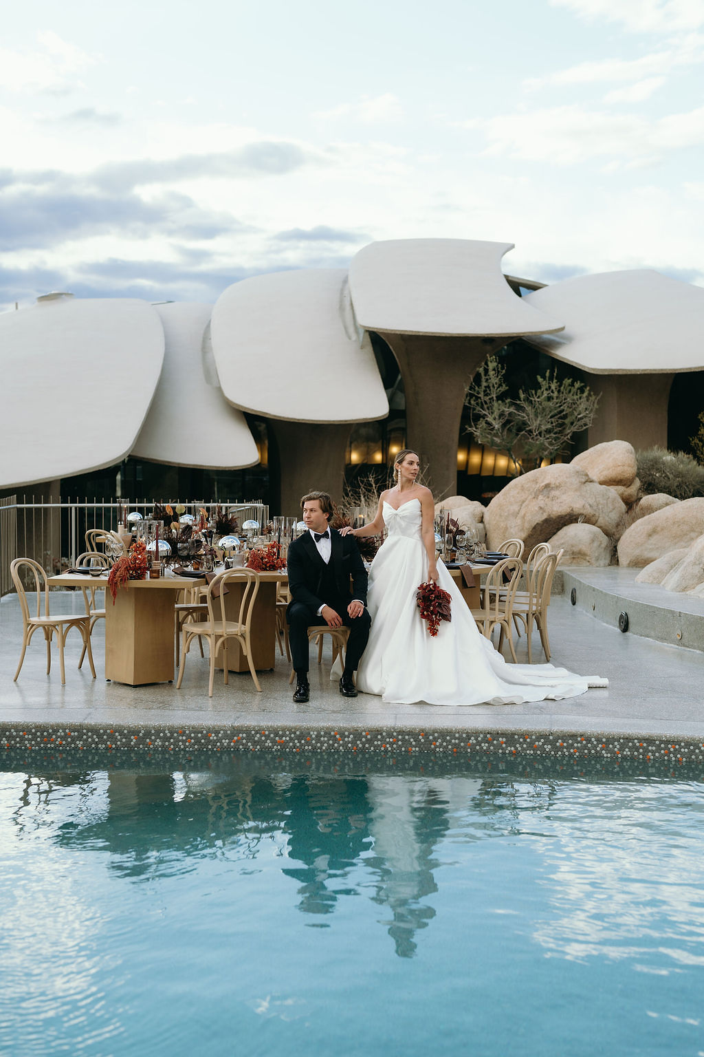 A bride and groom pose by a pool with modern architecture and outdoor dining tables in the background under a partly cloudy sky.