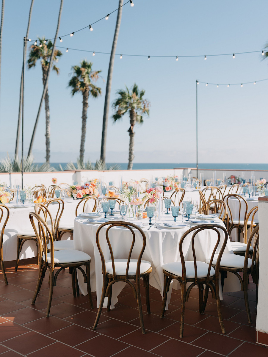 Outdoor event setup with round tables covered in white linens, wooden chairs, floral centerpieces, string lights, and palm trees in the background by the ocean.