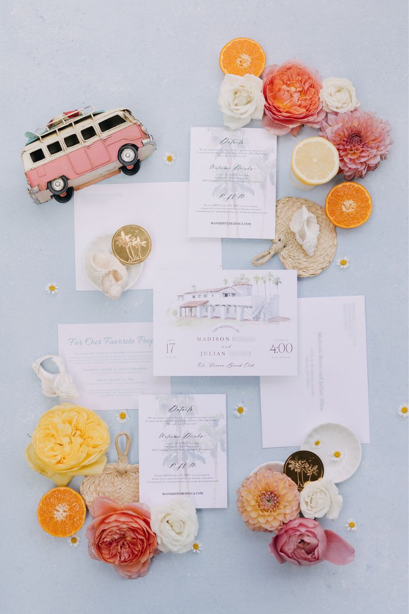 Flat lay of wedding invitations, pastel envelopes, colorful flowers, citrus slices, and a pink van decoration arranged on a light blue surface.