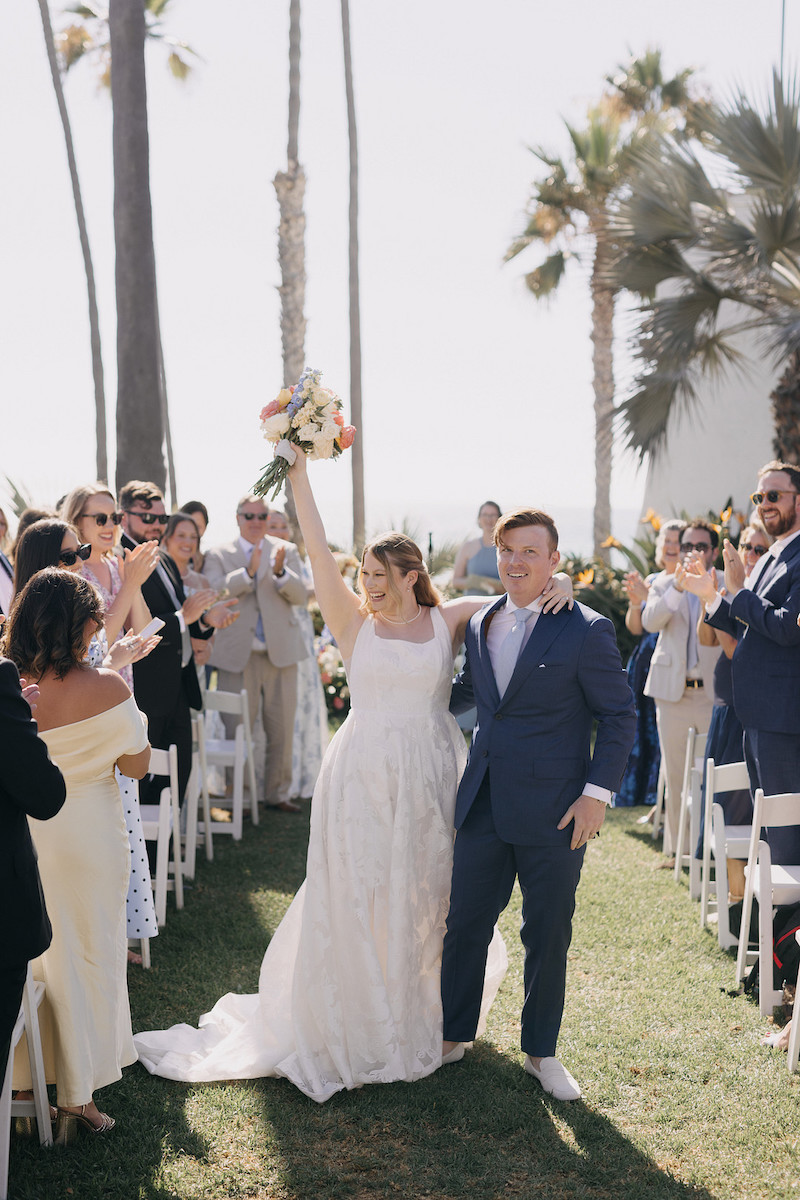 A newlywed couple walks down an outdoor aisle, smiling as guests stand and clap. The bride holds a bouquet aloft. Palm trees are visible in the background.