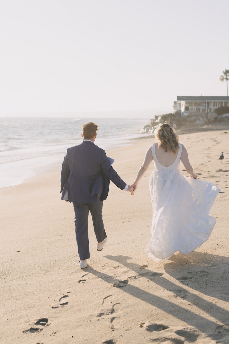 A couple in wedding attire walks barefoot, holding hands along a sandy beach with the ocean and buildings in the background.