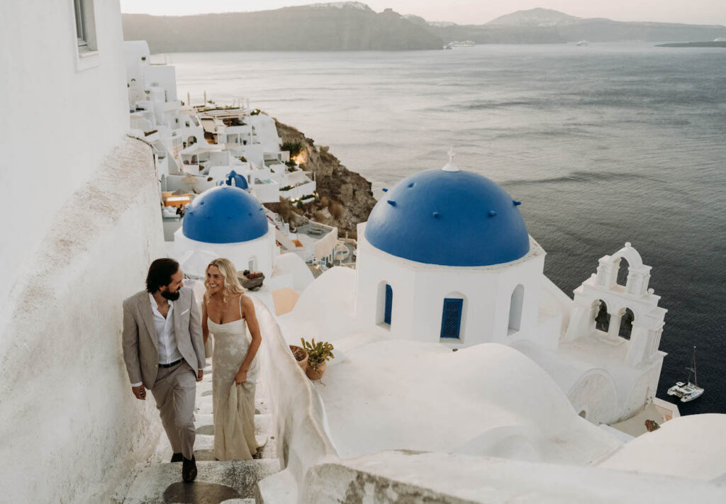 A couple in formal attire walks up white stairs beside blue-domed buildings, overlooking the sea in Santorini, Greece, at sunset.