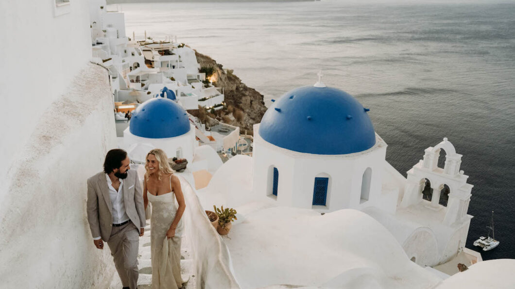 A couple in formal attire walks up white stairs beside blue-domed buildings, overlooking the sea in Santorini, Greece, at sunset.