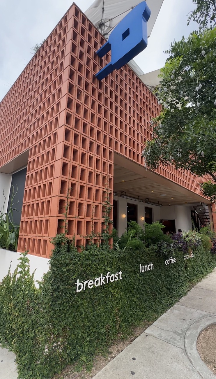 A modern red brick building with a blue geometric sign above a patio. The hedge in front displays the words “breakfast lunch dinner” in white letters.