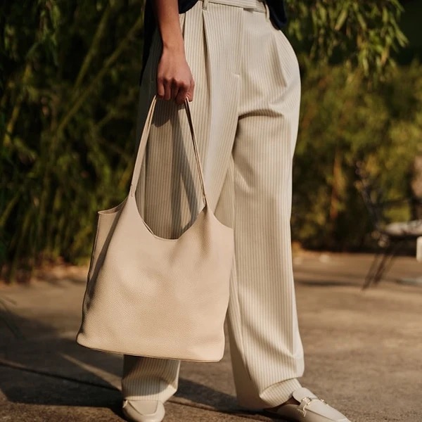 Person wearing light-colored pleated pants and white shoes standing outdoors, holding a large beige tote bag by their side.