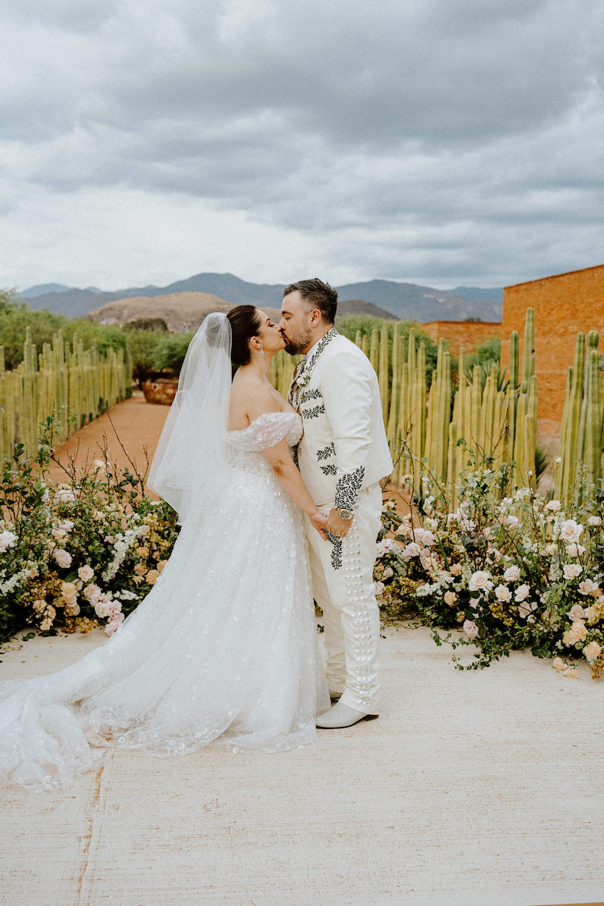 A bride and groom in wedding attire stand facing each other and kiss outdoors, with flowers, tall cacti, and mountains in the background under a cloudy sky.