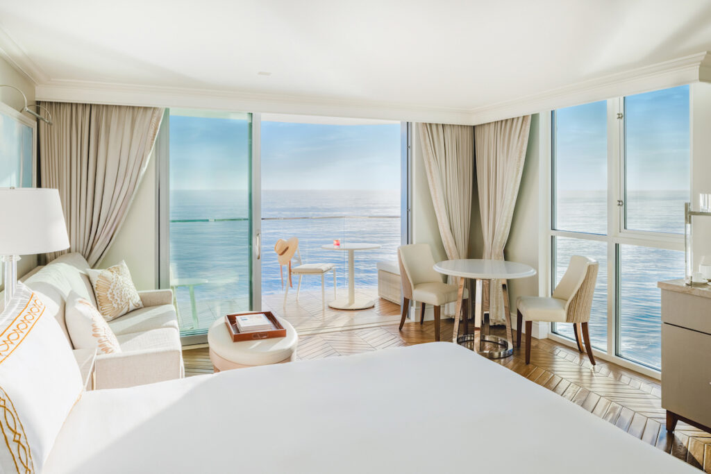 Bright hotel room with large windows, ocean view, bed, sofa, table, chairs, and balcony with a small table and red drink.