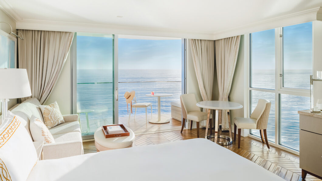 Bright hotel room with large windows, ocean view, bed, sofa, table, chairs, and balcony with a small table and red drink.
