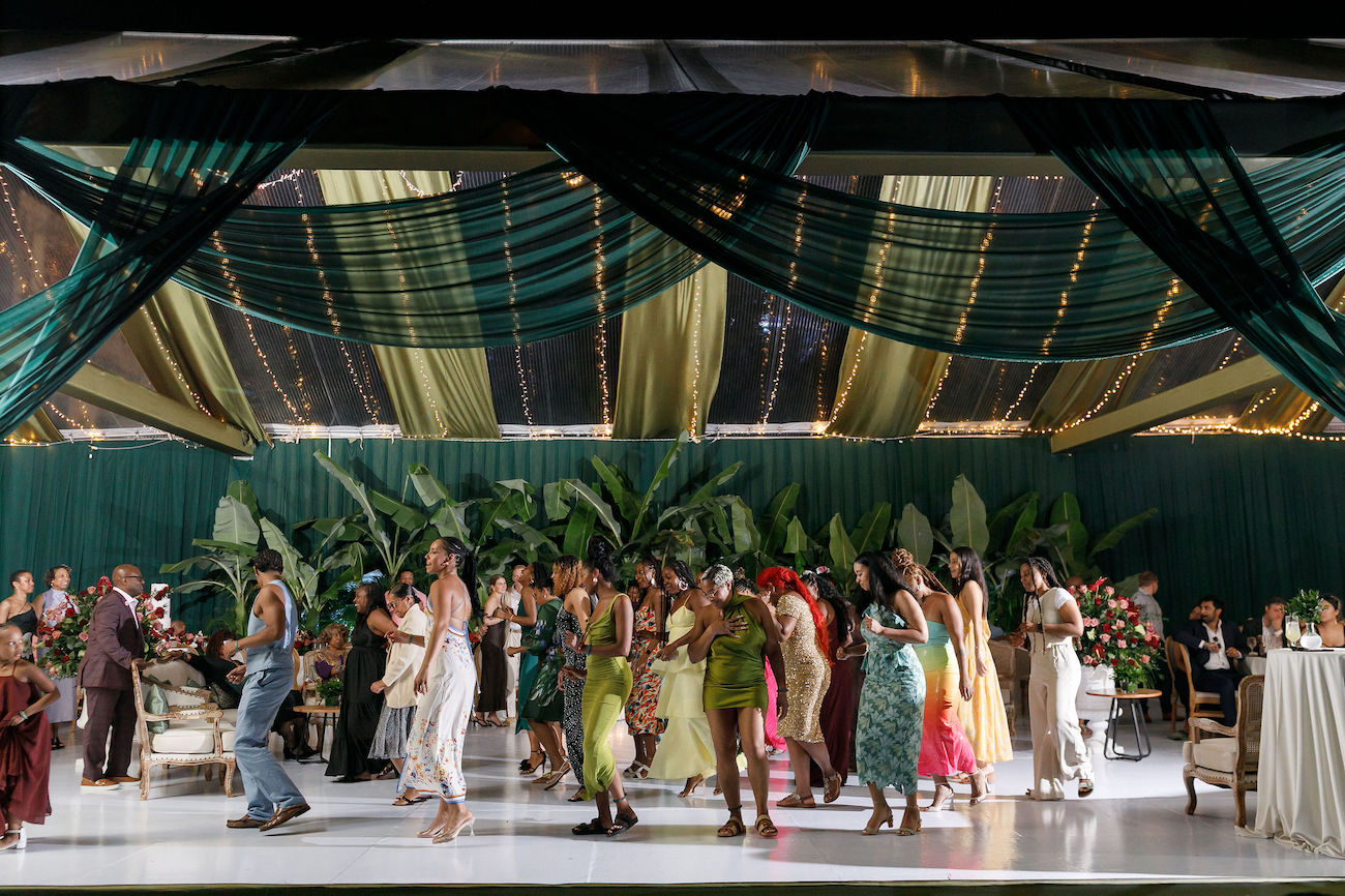 A group of people in colorful attire socialize and dance under a draped canopy with green plants and elegant decorations.