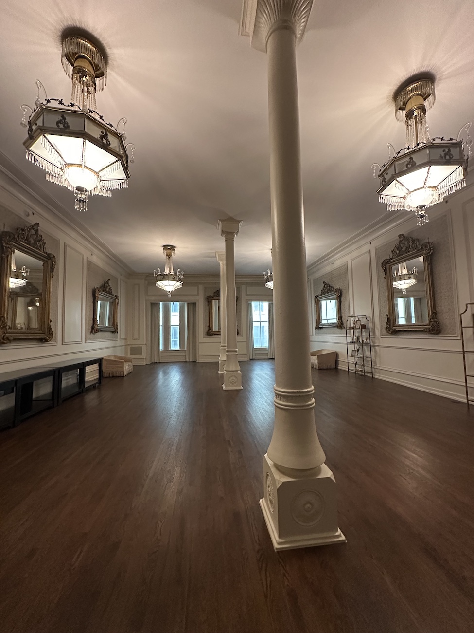 Spacious room with wooden floor, ornate mirrors on white walls, large pillars, and decorative ceiling lights.