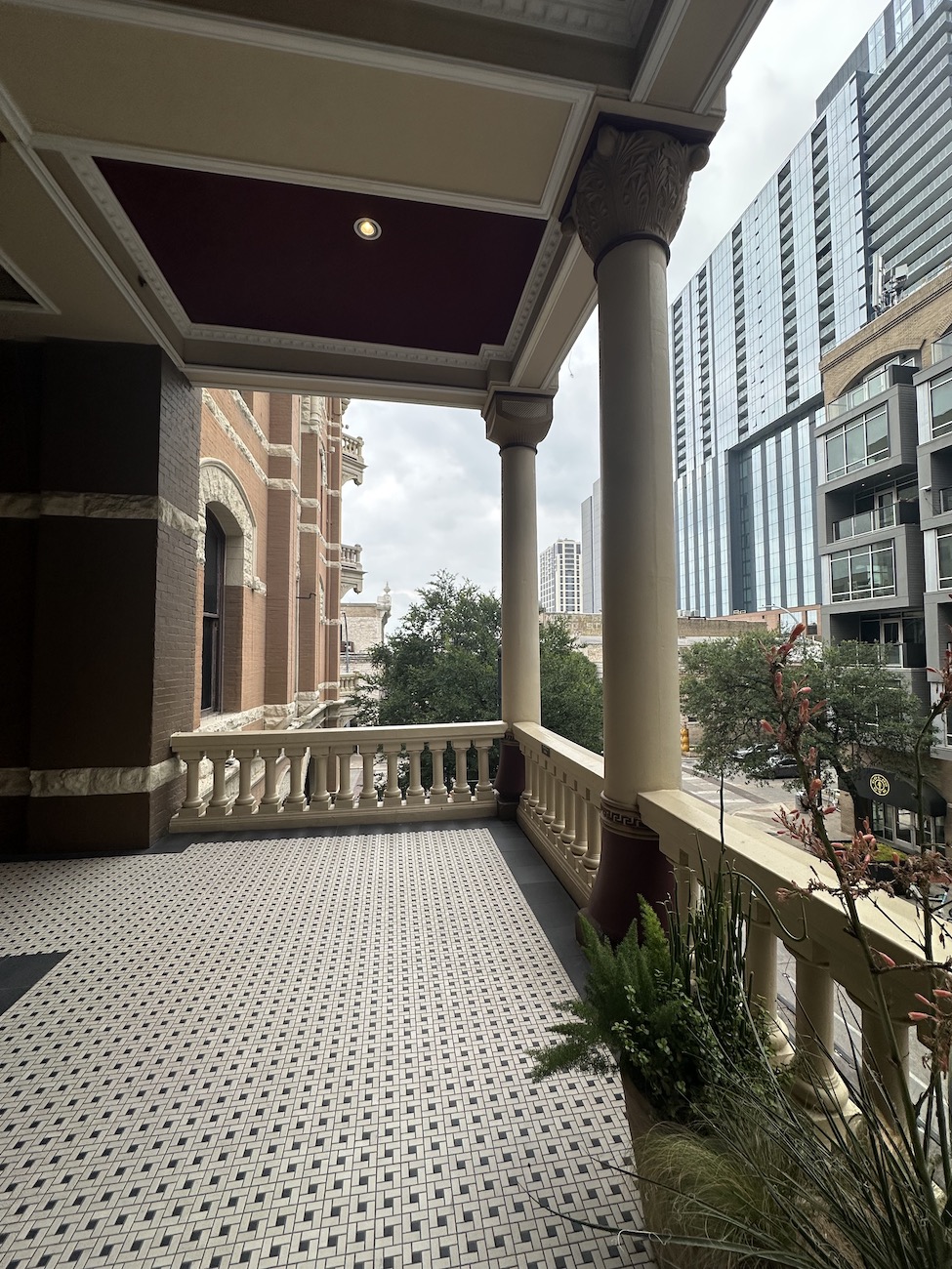 Covered balcony with patterned tile floor, columns, and city buildings visible in the background on a cloudy day.