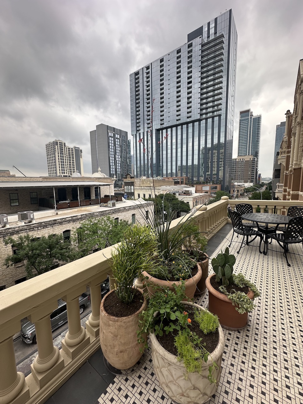 Potted plants on a balcony with a patterned tile floor overlook a cityscape of tall modern buildings under a cloudy sky.