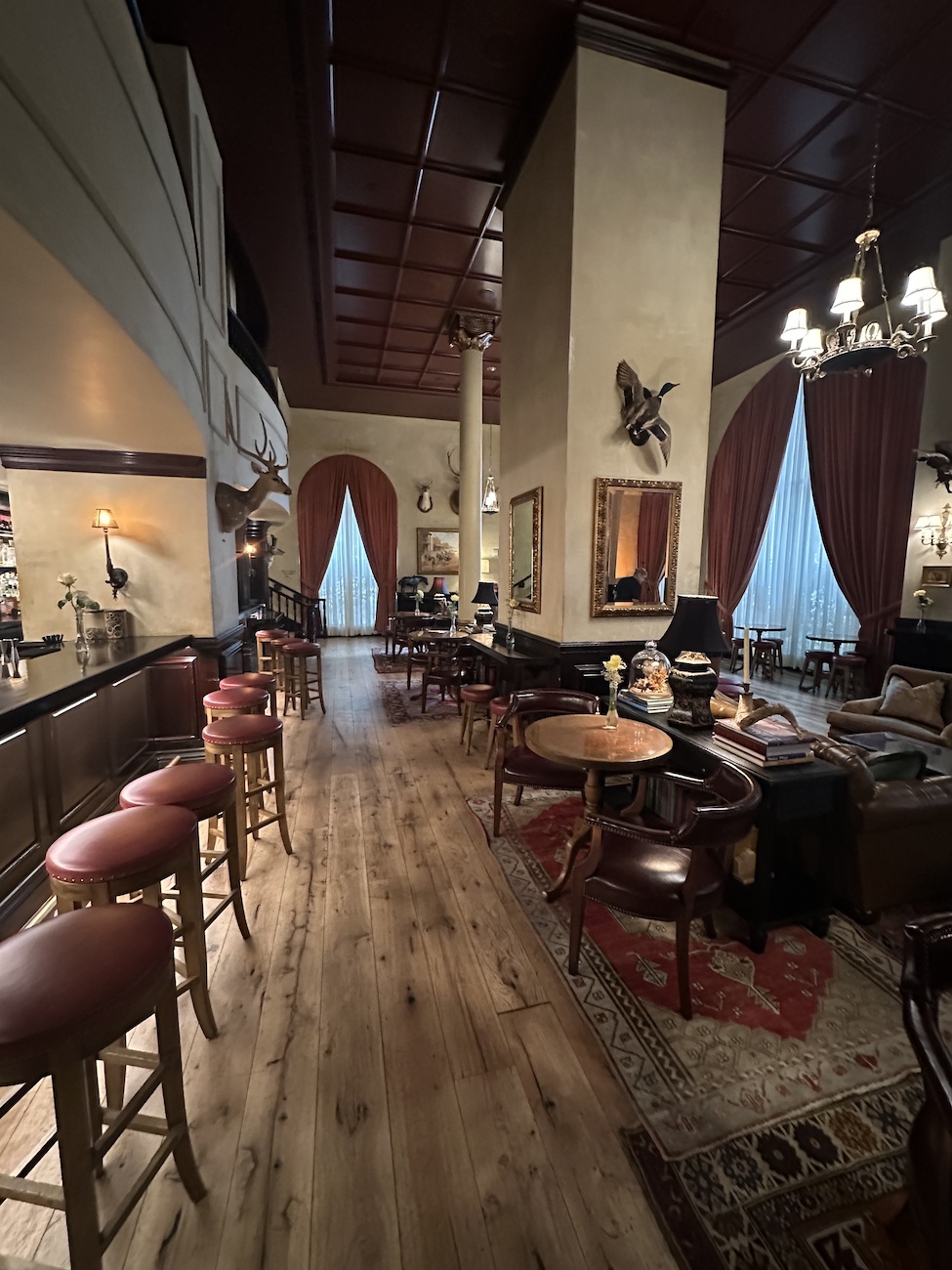 A dimly lit lounge with wooden floors, round tables, leather chairs, a bar on the left, and tall windows with red curtains.