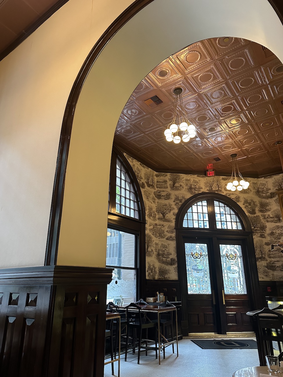A restaurant interior with arched doorways, stone walls, wooden trim, and a copper-colored patterned ceiling. Tables and chairs are arranged near large windows and a glass door.