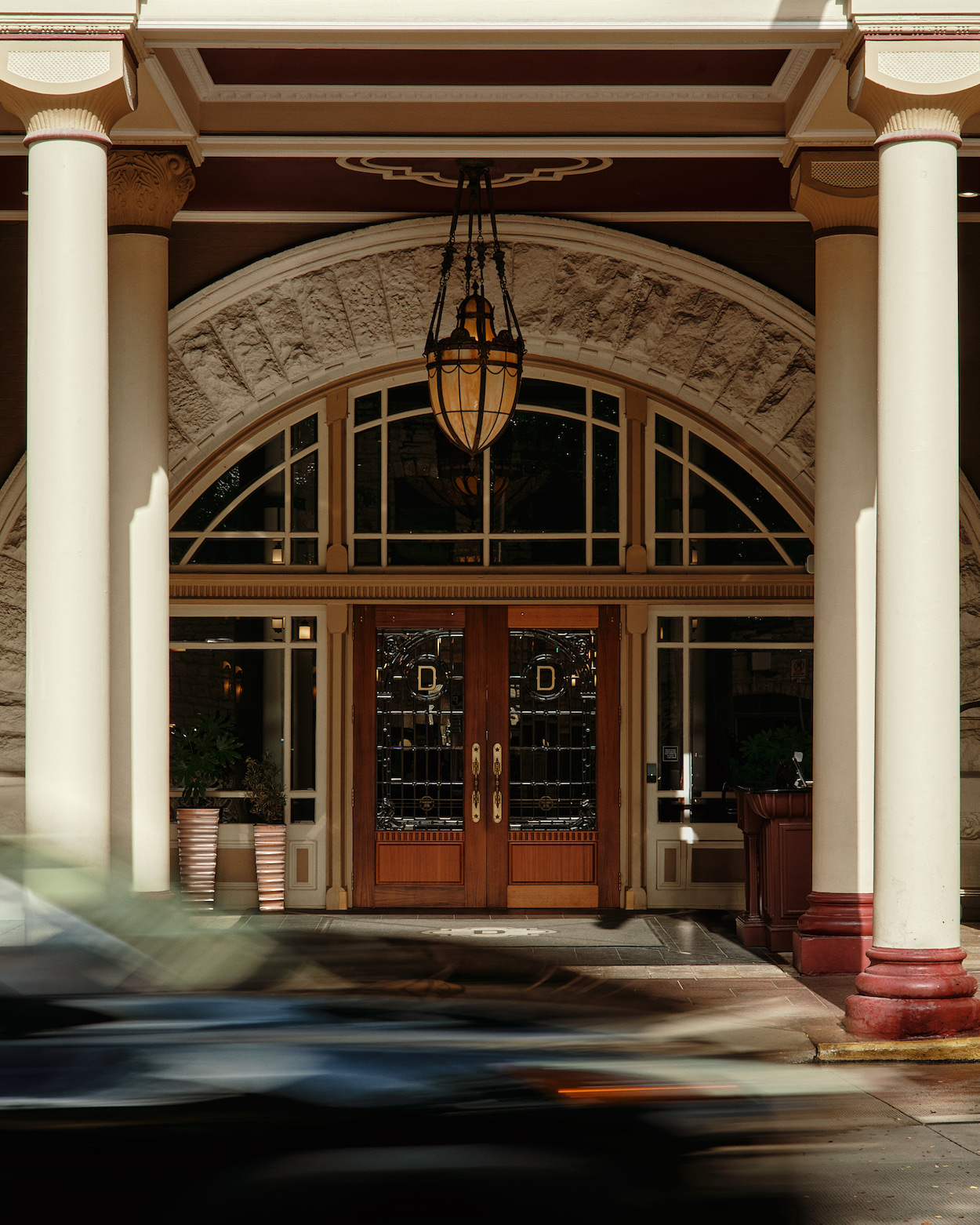 Ornate entrance of a building with glass doors, arched windows, white columns, and a large hanging lantern overhead. A blurred car passes by in the foreground.