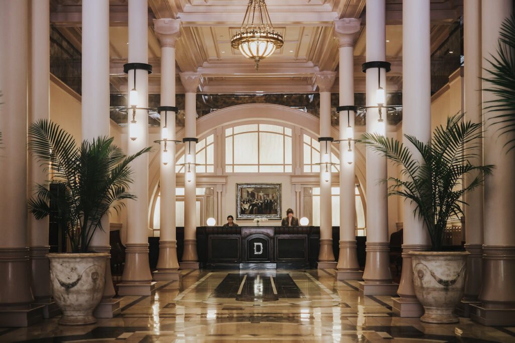 Hotel lobby with marble floors, tall white columns, a chandelier, two palm plants, and two people standing behind a black reception desk.