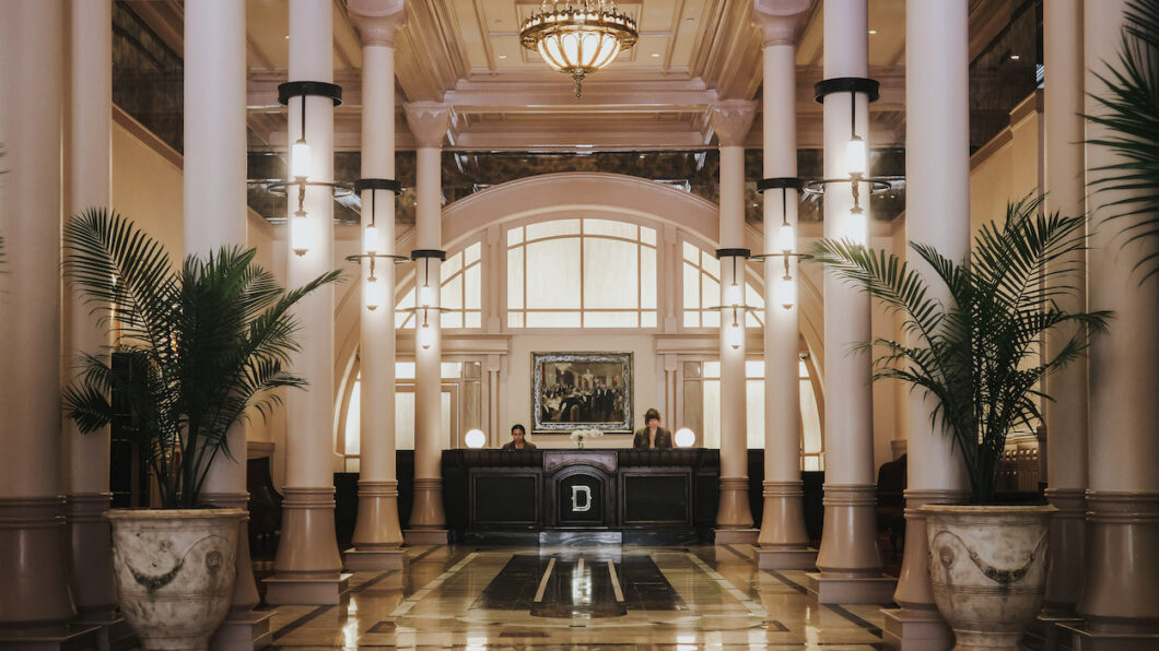 Hotel lobby with marble floors, tall white columns, a chandelier, two palm plants, and two people standing behind a black reception desk.
