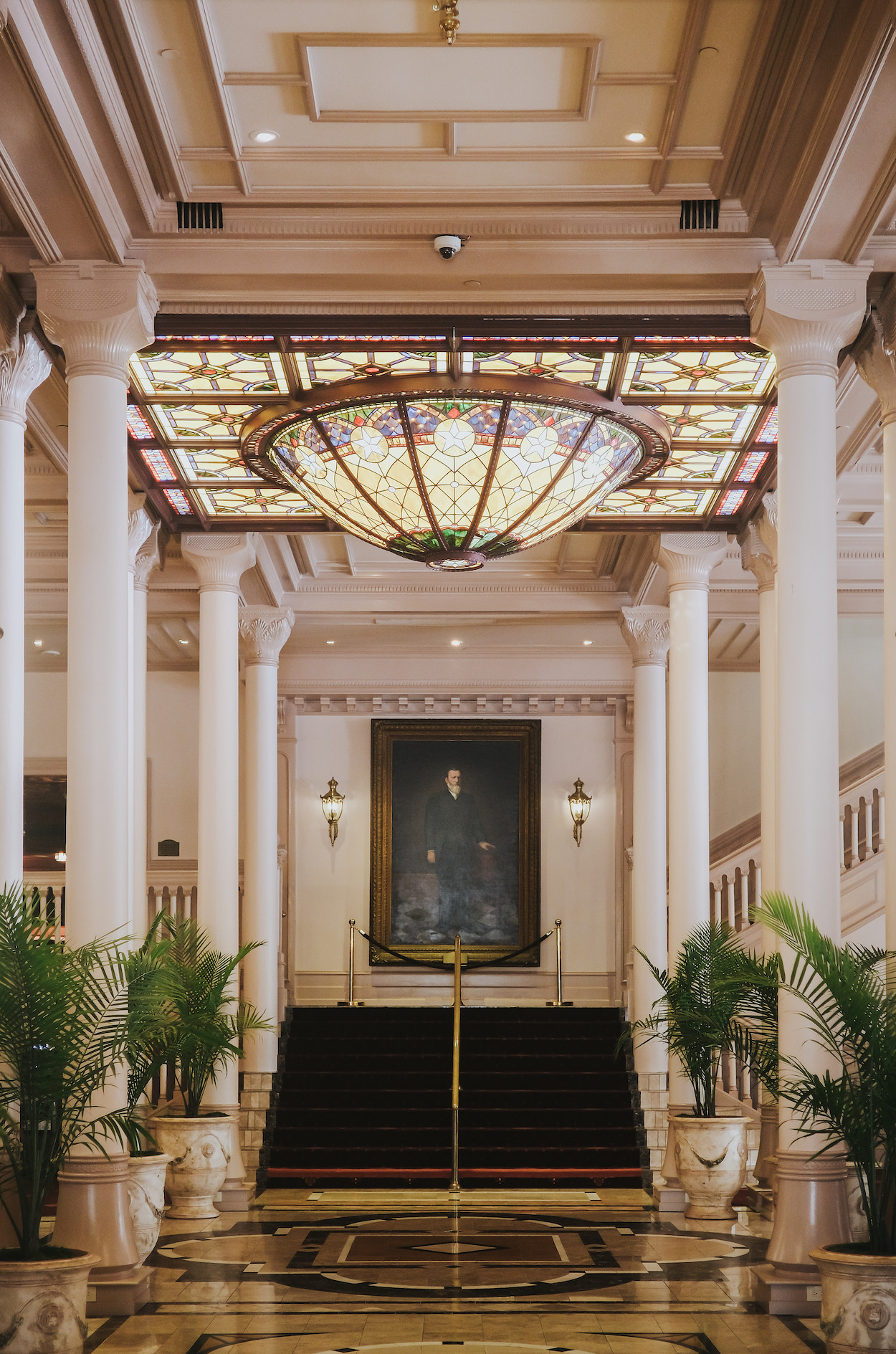Grand hotel lobby with tall white columns, stained glass ceiling fixture, red-carpeted staircase, a large portrait, and potted plants lining the entrance.
