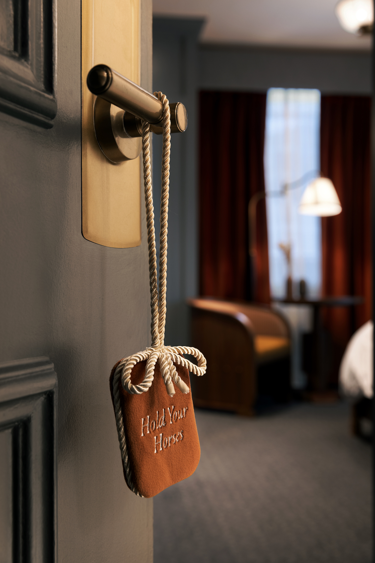 A hotel room door with a “Hold You Here” sign hanging from the handle; a chair, lamp, and window with closed curtains are visible in the background.