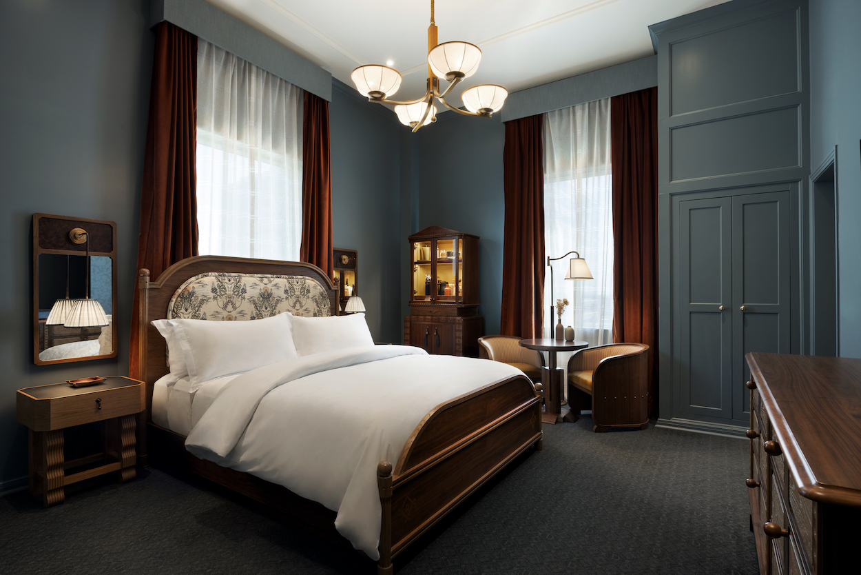 A hotel room with a large bed, two tall windows with curtains, wooden furniture, a chandelier, and soft lighting.