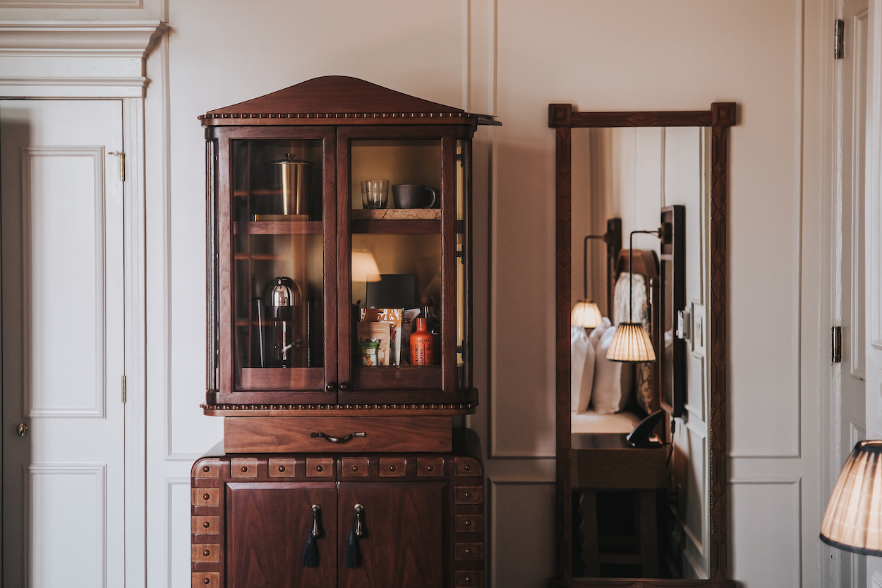 A wooden cabinet with glass doors is placed next to a tall mirror reflecting a lit lamp and part of a room.