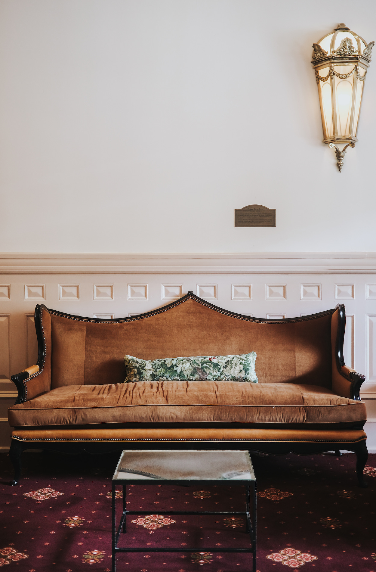 Vintage brown sofa with a green patterned pillow sits against a white wall, beneath a brass sconce, with a small metal table in front.