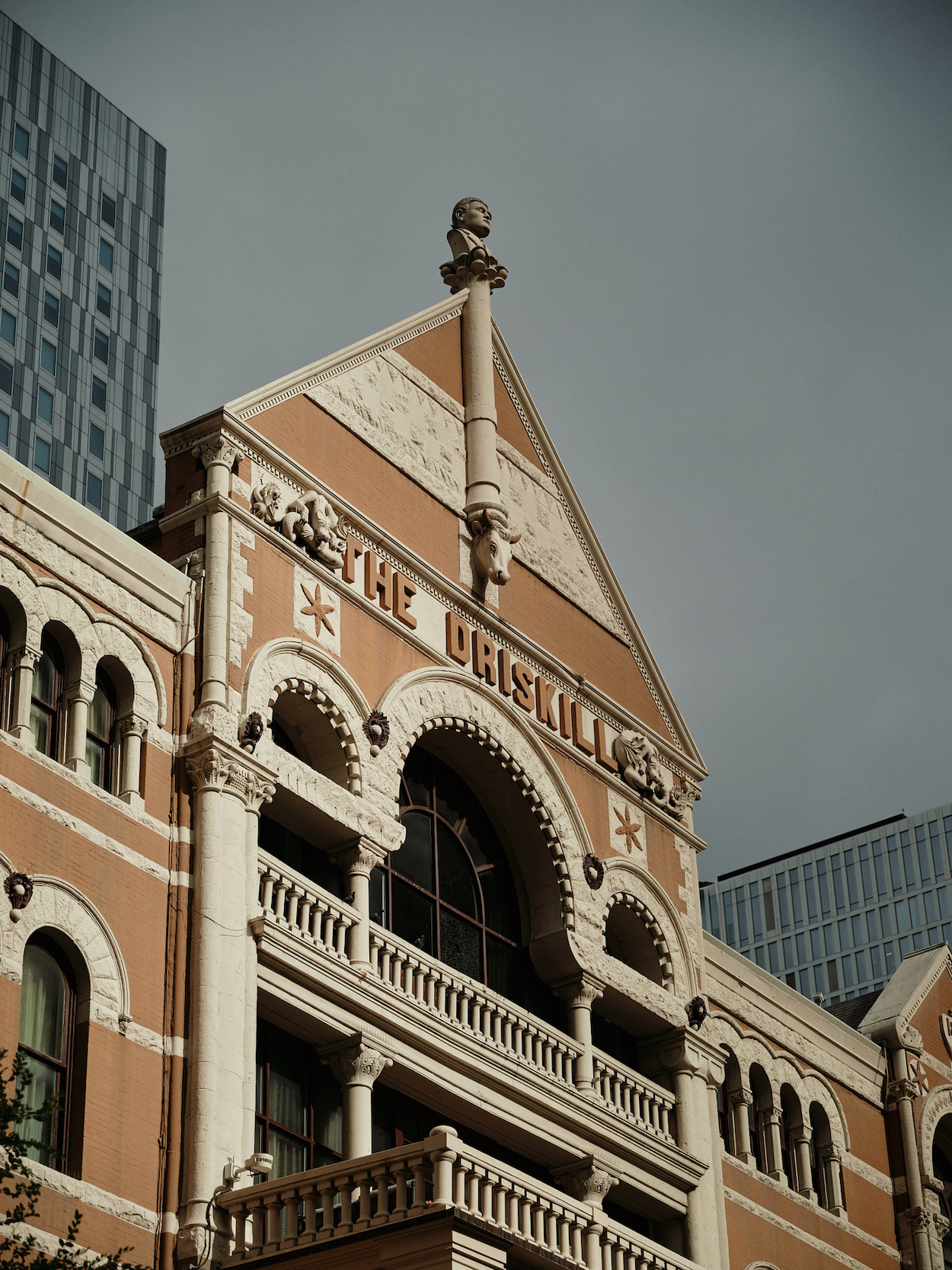 A historic building labeled “The Driskill” with ornate architectural details, arched windows, and balconies, set against a gray sky and modern skyscrapers.
