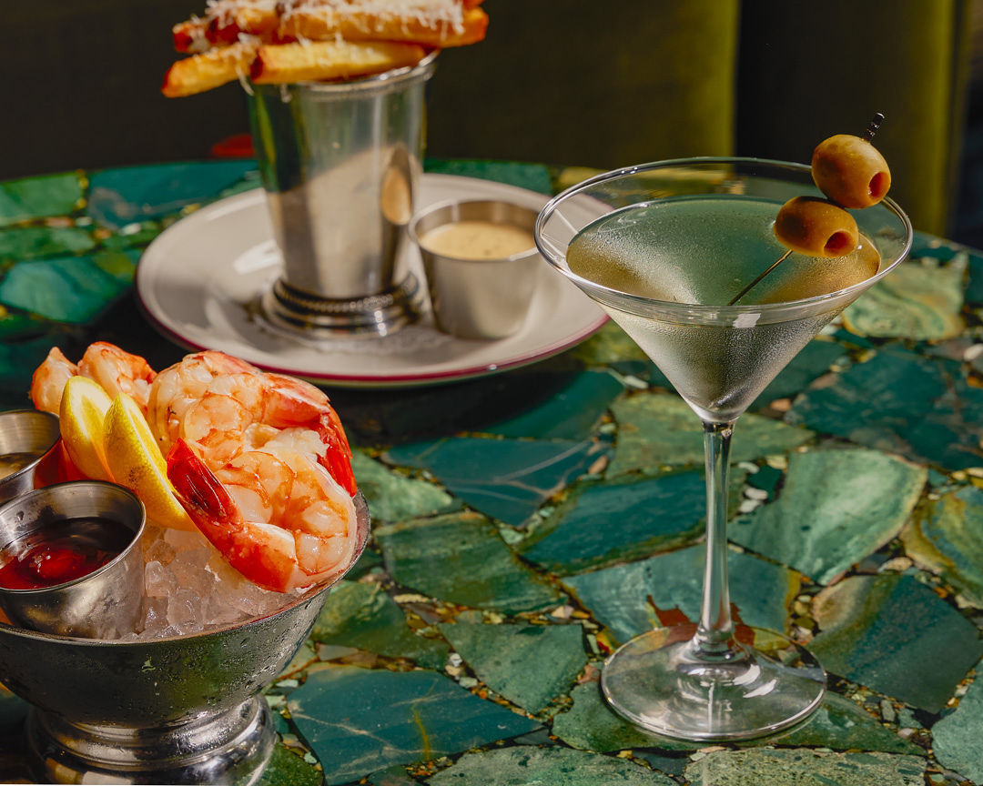 A martini with two olives, a bowl of shrimp cocktail, and a cup of fries with sauce are arranged on a green mosaic table.