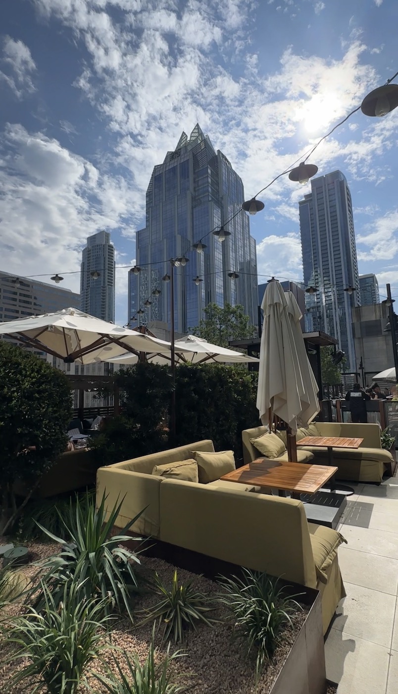 Outdoor patio seating with beige couches and umbrellas in front of the tall modern skyscrapers of Austin, Texas under a partly cloudy sky in an urban setting.