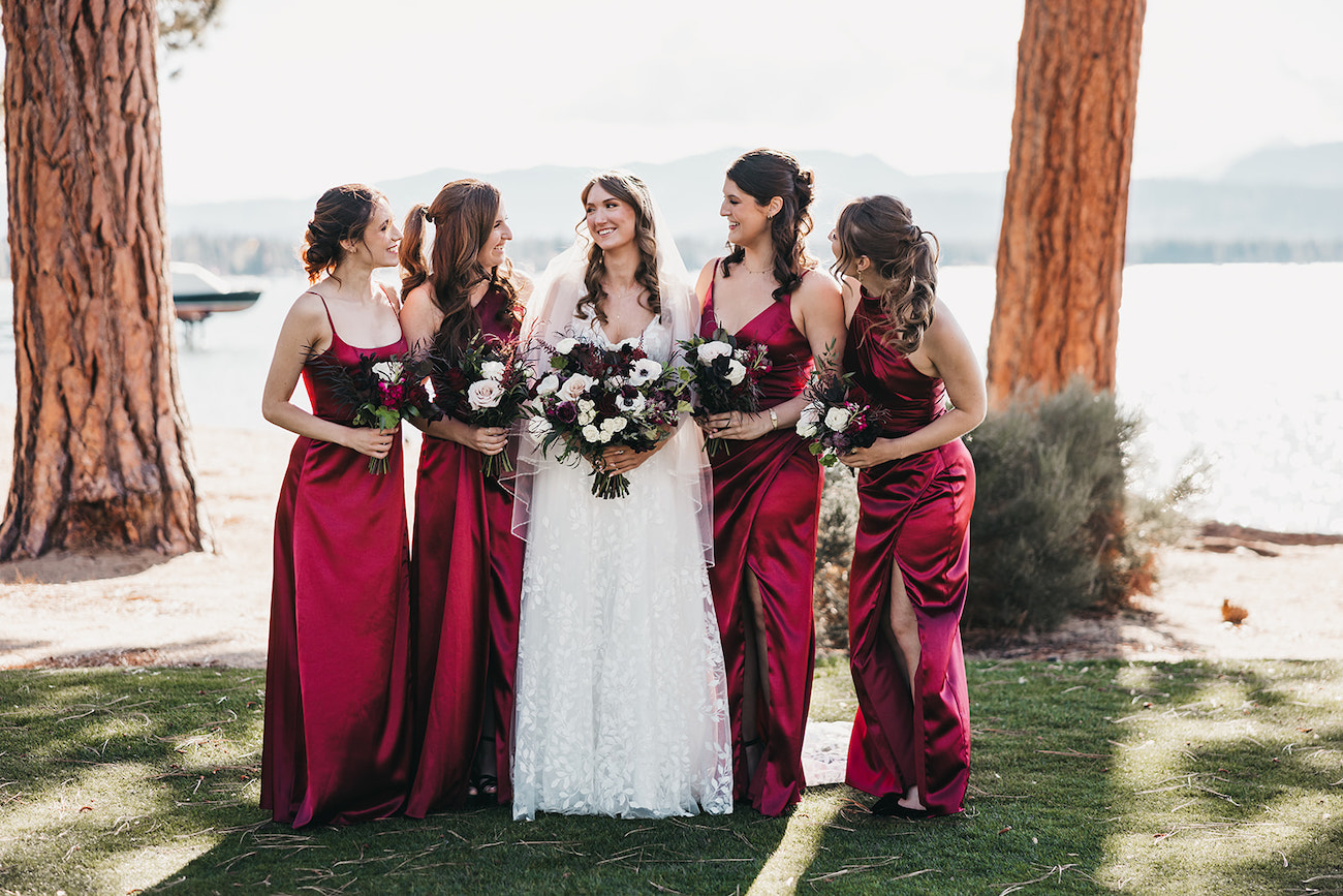 A bride in a white dress stands outdoors with four bridesmaids in matching red dresses, all holding bouquets, near trees and a body of water.