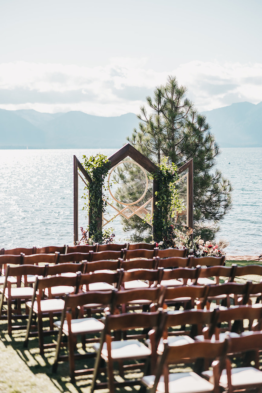Rows of empty wooden chairs face a simple wooden wedding arch decorated with greenery, set by a lake with mountains and trees in the background on a sunny day.