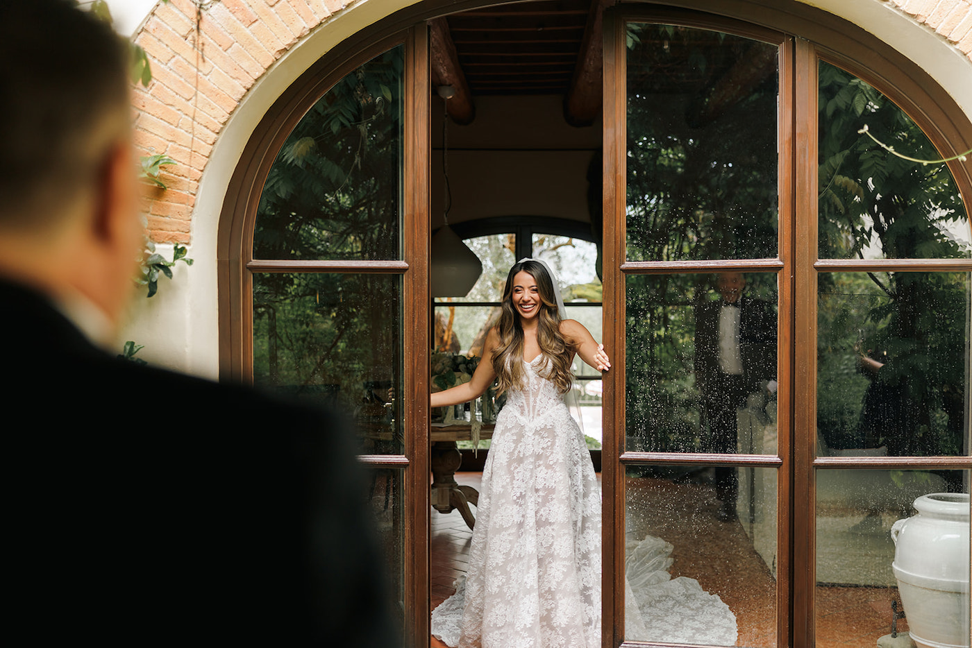 A woman in a white dress stands smiling behind a glass door, facing a person in a suit in the foreground.