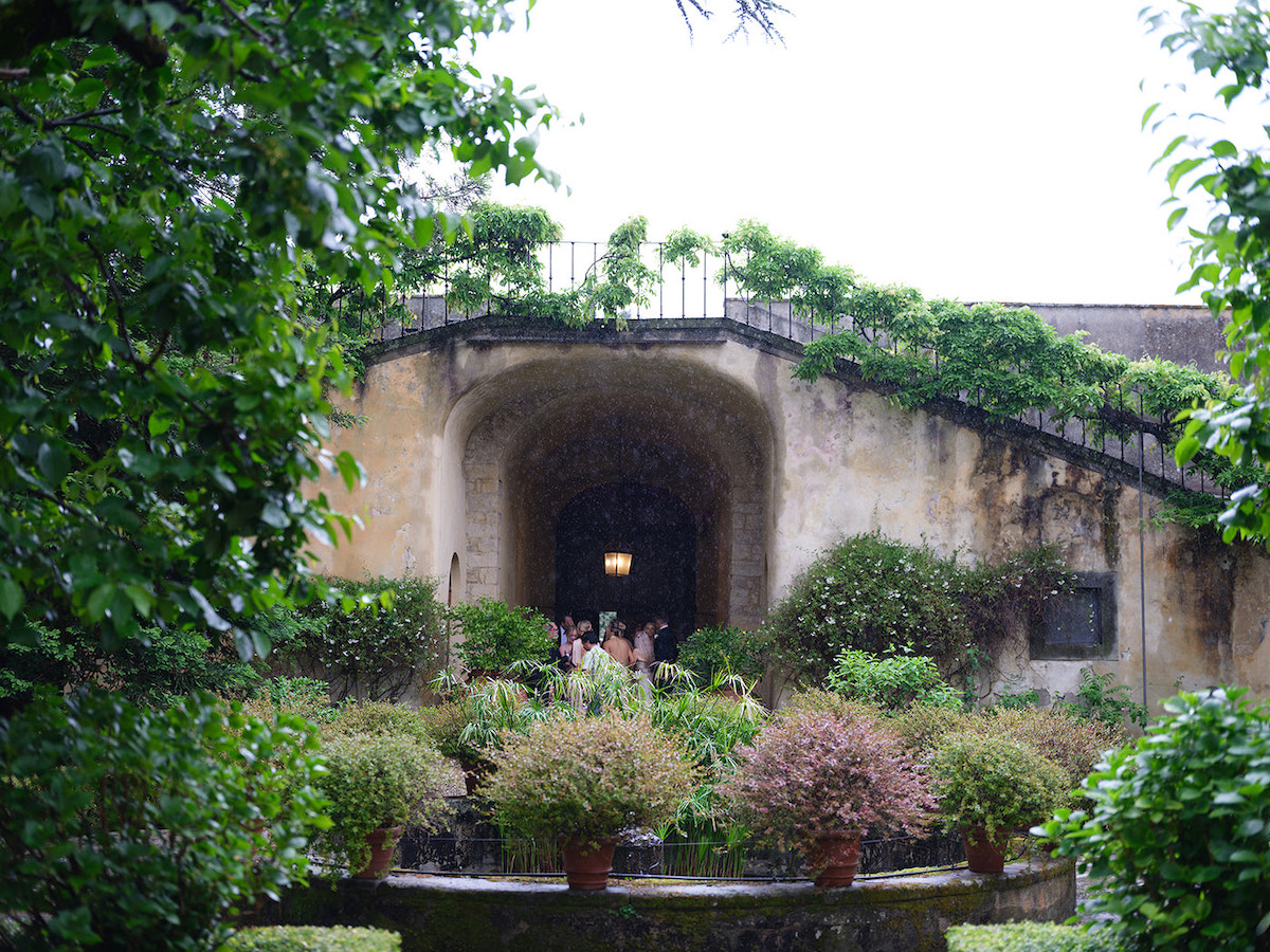 Stone building with arched entrance surrounded by lush greenery, potted plants, and shrubs; people visible near the doorway under an overcast sky.