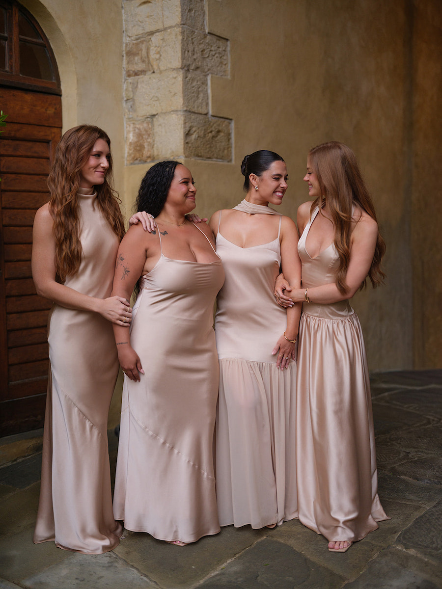 Four women in light beige dresses stand together indoors on a stone floor, smiling and holding each other’s arms in front of a stone and wood wall.