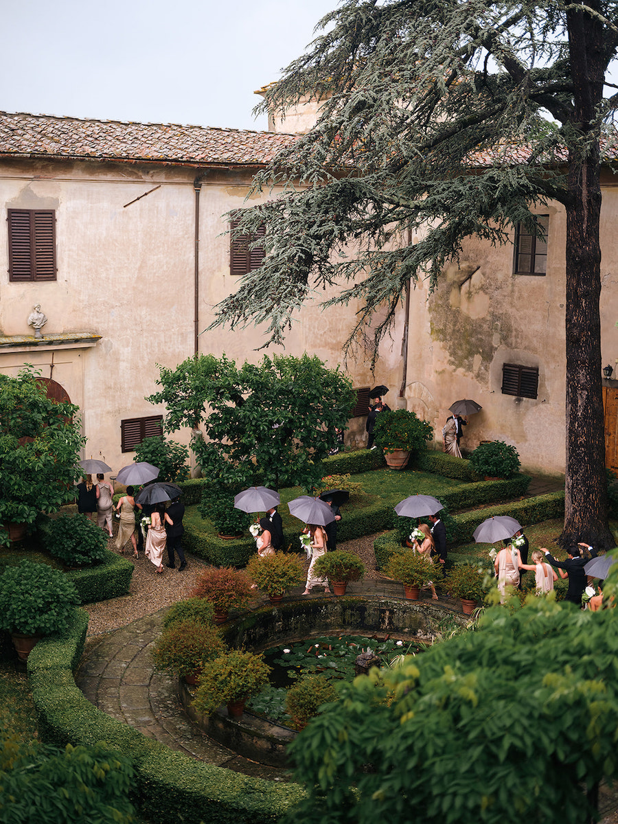 People holding black umbrellas walk through a formal garden with trimmed hedges and a fountain, next to an old stone building with shuttered windows.