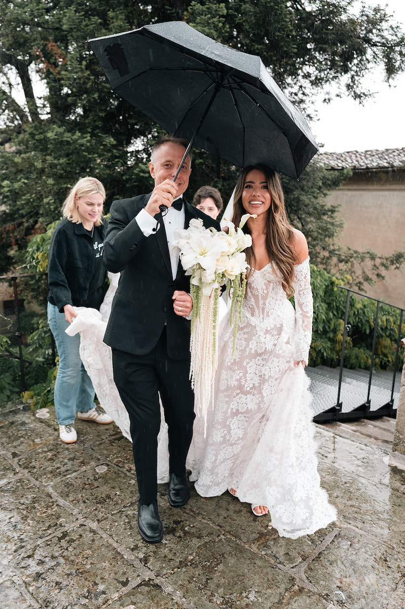 A bride in a white lace gown walks outside on wet pavement under a black umbrella held by her father in a suit, with two women assisting.