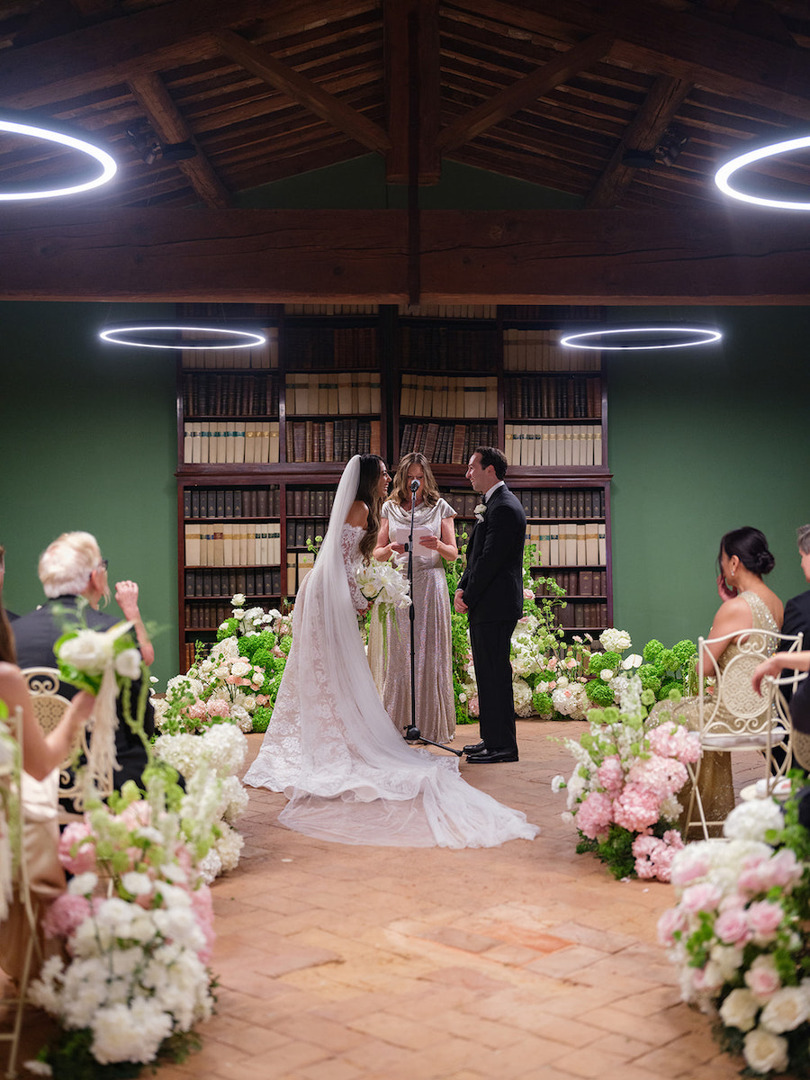 A bride and groom stand with an officiant during an indoor wedding ceremony, surrounded by floral arrangements and seated guests.